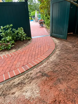 Curved red brick walkway leading through a garden toward a green gate.